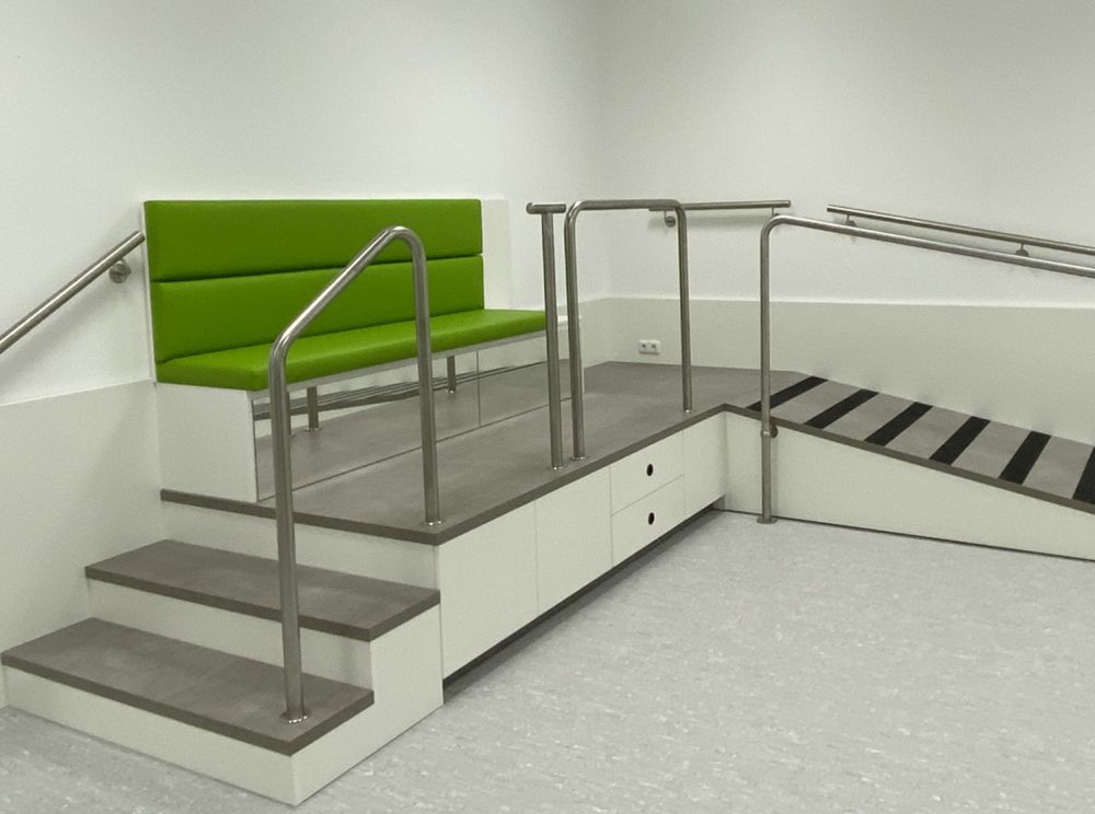 Maßgefertigtes Podest von Bäuerlein Objektdesign für den barrierefreien Zugang zur Anprobe orthopädischer Hilfsmittel. Das Podest ist weiß, mit einer Lauffläche in Betonoptik und einem stabilen Geländer zur sicheren Nutzung. Die Polsterflächen sind in einem freundlichen Grünton gehalten und farblich auf das Gesamtkonzept des Sanitätshauses abgestimmt.